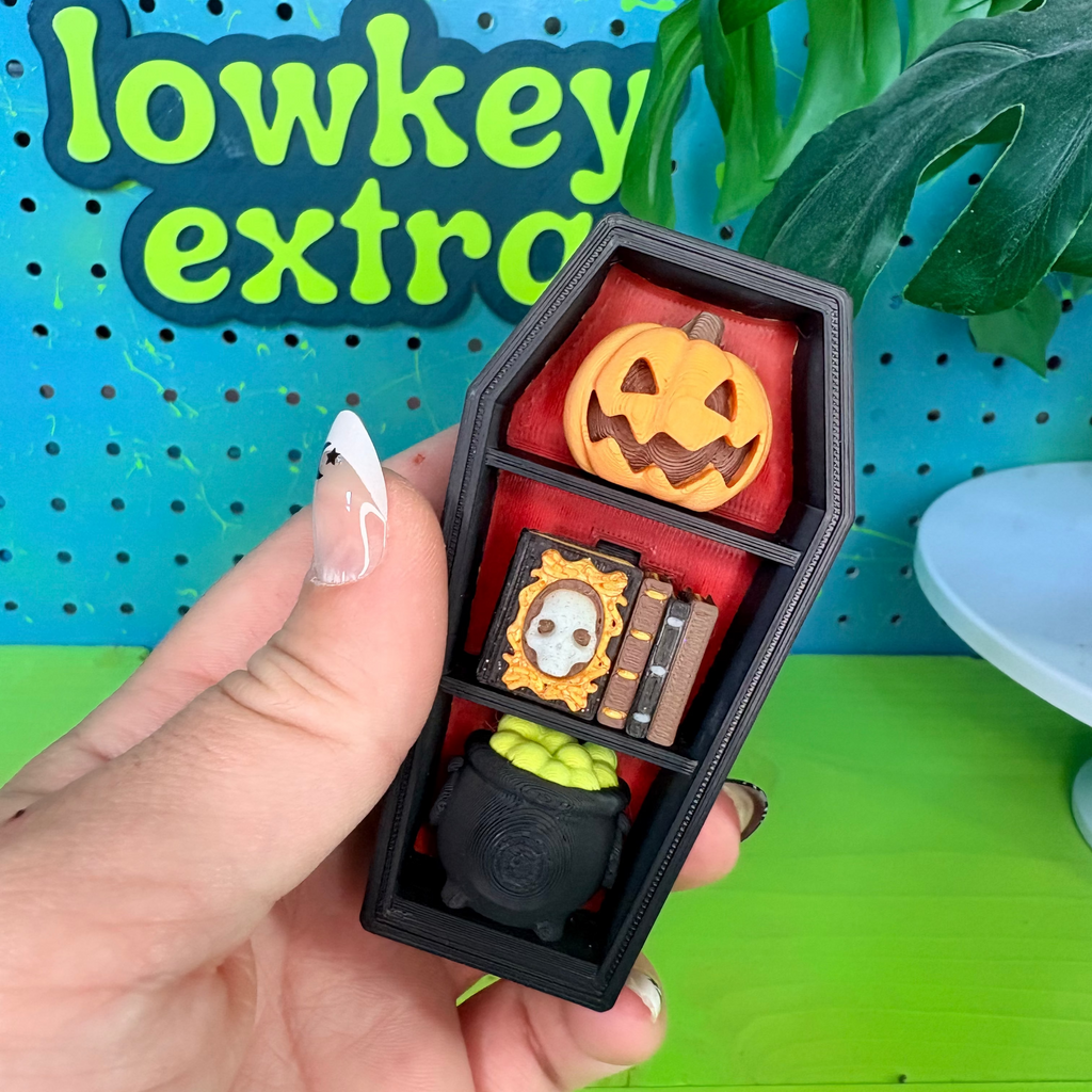 A hand holds a small black coffin-shaped organizer filled with Halloween-themed miniature accessories, including a jack-o'-lantern, a cauldron with green contents, and a small book with a skull illustration. The background features a bright blue pegboard with green text reading 'lowkey extra' and green foliage.
